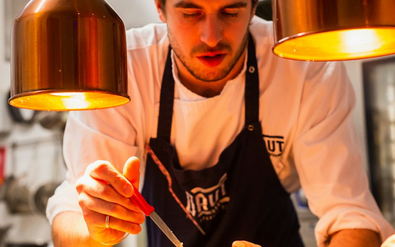 Chef plating a dish under heat lamps in a restaurant kitchen